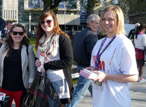 Two women taking flyers from a smiling woman in a white tee shirt with pink logos