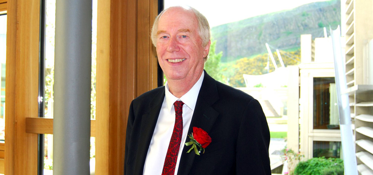 Malcolm Chisholm smiling with a red rose button hole with a view of the Scottish Parliament looking towards Salisbury Crags visible through the window behind him