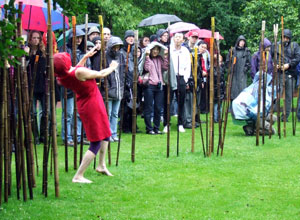Woman with a red head scarf covering her face and wearing a red dress, dances round the bamboo rods