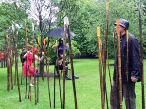 Woman with a red head scarf covering her face and wearing a red dress, dances round the bamboo rods, while Chinese musician in rain proofs blows, musically, on some of the bamboo rods