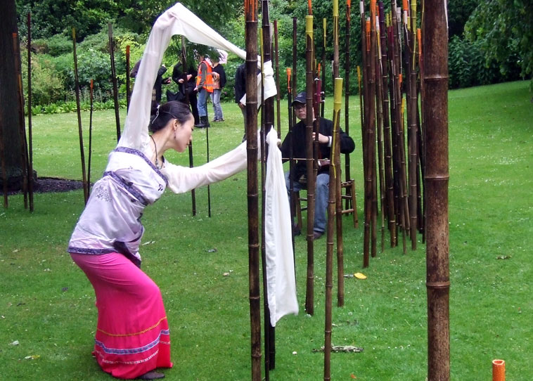 Dressed in white, lilac and a cerise skirt Chang Chang Zhang embraces the rods of Bamboo while a seated musician plays