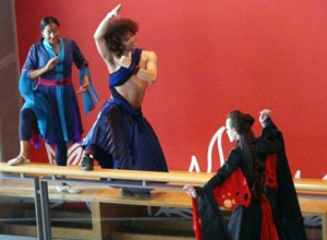 Two dancers stand on a banister against a red wall confronting a black and red caped stilt walker