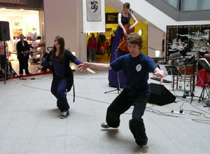 A young woman and a young man in Taichi poses as Louise Martin in a satin purple evening dress, stands on stilts and plays her cello