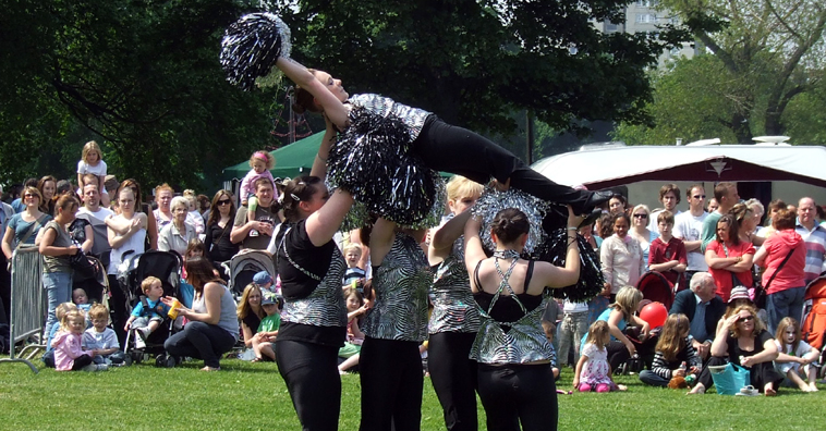 Young women in black slacks and silver tops shake silver and black pom poms, raising one of their team onto her back