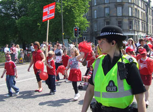 A policewoman in the foreground watches over pupils and staff in red, with red pom poms and balloons