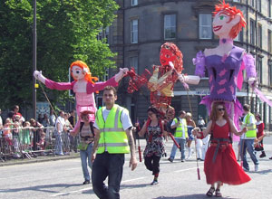 Three puppeteers carrying eight foot pink, red and violet clad puppets on their shoulders