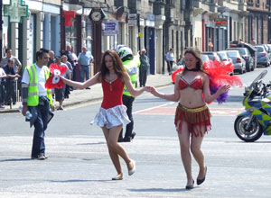 Two young women in red, samba in the middle of the street, as a worker and pollice watch on in flourescent yellow.