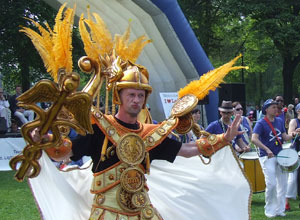 A gold and white clad man with golden feathers dancers with his caduceus as blue and white dressed men drum in the background