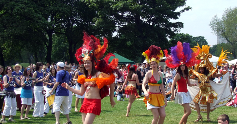Five brightly costumed dancers with the drummers in the background in blue t-shirts and white trousers