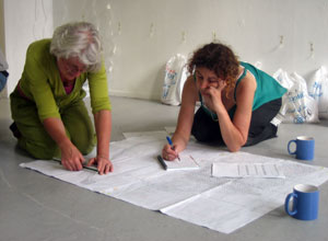 Jane and Ally kneeling over a giant sheet of paper planning the architecture of the installation with blue coffee musgs in the foreground