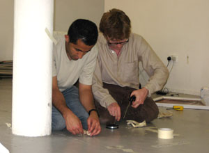 Two men kneeling, one tying a figure to a wire, the other measuring from a spool