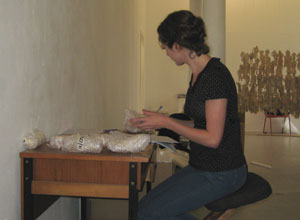 Woman seated at a desk covered with bubble wrapped figures as she lists the details for each figure, to make sure they find the ir right place in the next installation