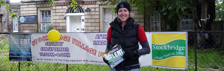 Lorna Cameron smiling in black sleeveless puffer jacket in front of the Stockbridge Village Festival banner hanging outside the Braeburn House Hotel  