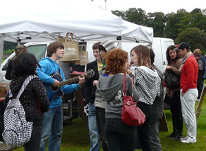 Young man in blue with a guitar singing to a small group of young people standing in front of The Store stall