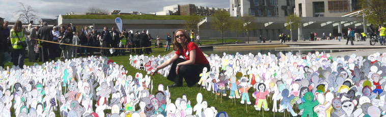 Claire Aston (in a red Christian AId tee shirt,) crouches at the heart of the Saltire of paper figures with the terraced of the Scottish Parliament in view behind her