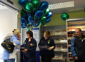 A woman takes a biscuit from two women members of staff with green and blue helium balloons in the background in Scotmid colours