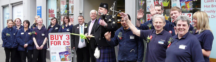 Store manager, Billy Stirling and staff standing outside Easter Rd Scotmid, smiling and waving, with Mark Lazarowicz and a piper, ready for the opening ceremony