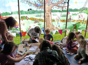 Adults and children working away at arts and crafts with a painted tree on the canvas of the tent
