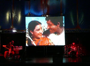 Big screen showing  wife and husband smiling at  each other as they work the land, with  musicians lit red below.