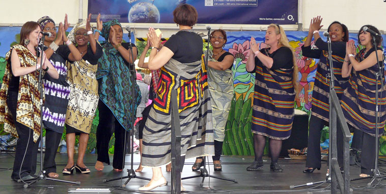 Eight women in African dress singing, clapping and dancing led by a ninth woman facing them