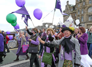 Women marching down North Bridge with purple flags and purple and green balloons