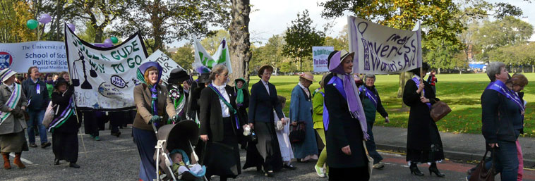 People in Edwardian dress marching with banners including banners "We've a gude way to go", and Inverness Women's Aid