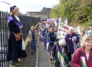 A woman dressed as a suffragette stands by the railings as people march up the hill with the old Scottish Office in the background