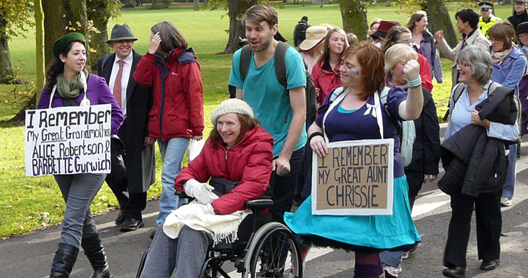 Smiling woman with a white  knitted beret in a wheelchair with two young women carrying placards: "I remember my great Grandmothers" and "I rememberr my great aunt Chrissie"