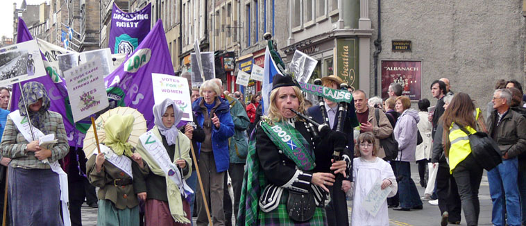 Piper in green and blue tartan leads a procession of women in children dressed in Edwardian costume down the Royal Mile