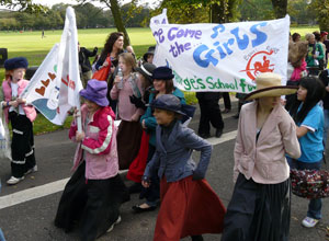 Pupils in Edwardian era hats carrying a banner "Here Come the Girls"