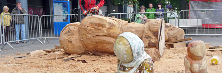 Seven foot tree trunk lying in a pile of sawdust with Arran Ross' chain saw carving it as Leithers watch on