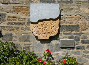 Grey and sand coloured bas relief sculpture on garden wall with red roses beneath