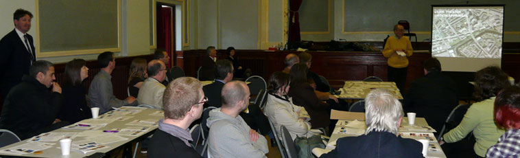 View of  people sitting round tables looking towards Les making a presentation in the  Thomas Morton Hall, Leith