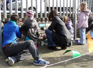 Three small children, a man and a woman painting bricks on the wall which is eight  bricks high