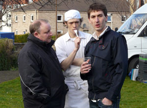 Three men - one in chef;s whites with a plastic fork in his mouth - conferring