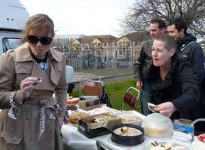 Woman in a raincoat and shades sampling a cake as Jane from Renroc watches on
