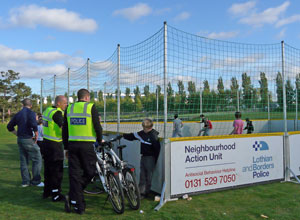 Two uniformed officers talk to a boy with a bicycle as boys play football in the netted and boarded moveable pitch