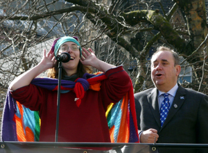 Penny Stone in red sweater and rainbow coloured flag raises her hands to her ears as Alex Salmond sings behind her