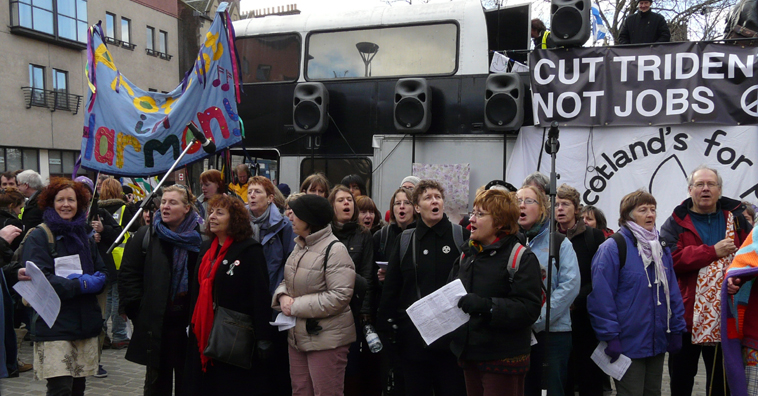 Protest in Harmony -  a couple dozen people singing with the double decker speakers; bus in the background with the banners CUT TRIDENT NOT FOR JOBS and Scotland's for Peace