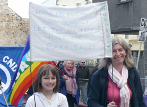 Girl and woman with banner "SPEND THE MONEY ON SCHOOLS. LET THE MILITARY HAVE THE BAKE SALES