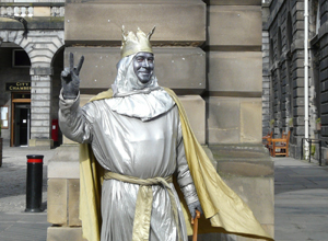 Man covered in silver paint with gold crown and cape posing as a statue outside the City Chambers and raising his fingers in the peace sign