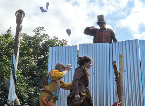 Man in C19th military costume standing behind corrugated iron wall throwing papers at woman and man mockingly