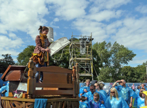 Woman on a boat with a  model of a crescent moon, surrounded by people in blue disposable ponchos with a blue clouded sky and trees behind her