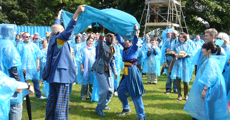 Audience in blue plastic ponchos surround three performers fishing under a blue sheet