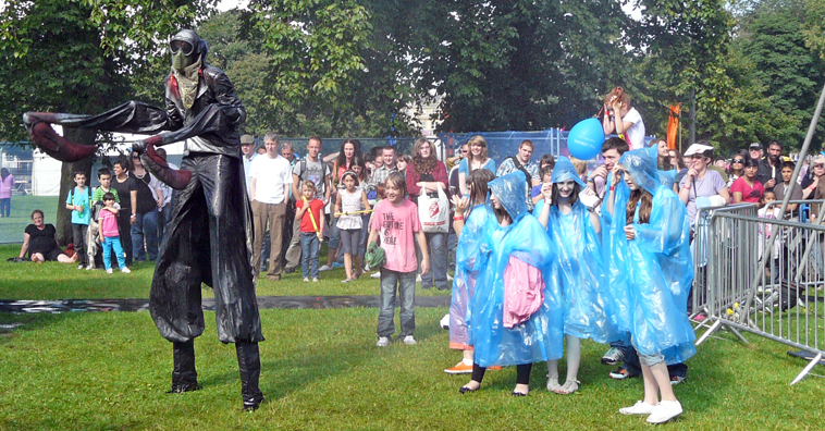 Audience in blue plastic ponchos surround three performers fishing under a blue sheet