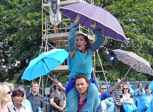 Performer with a purple umbrella being carried through the audience on a man's shoulders