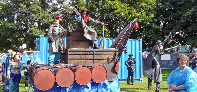 Pirate ship led by a stilted man in a scary praying mantis costume with pirates  posing and scowling