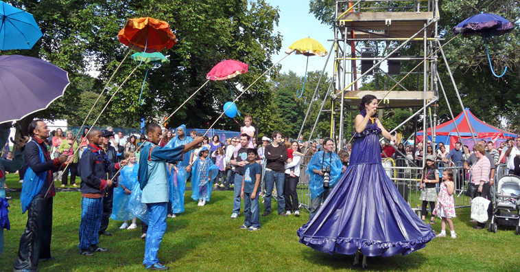 Performers holding brightly coloured umbrellas on poles with woman on stilts in purple dress and purple umbrella