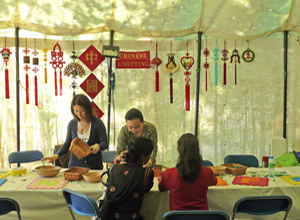 Two women demonstrating Chinese knotting craft with examples hanging on the tent wall behind them