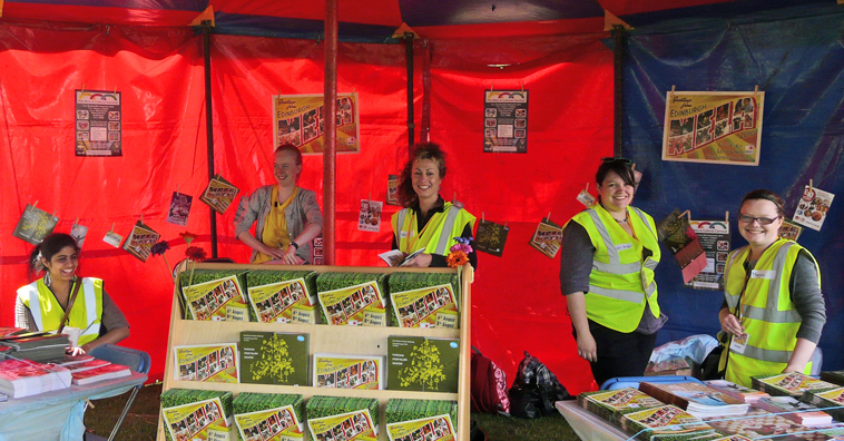 Five young women in yellow flourescent  jackets smiling in the red Mela 2010 information tent with the Mela 2010 postcard brochures on display